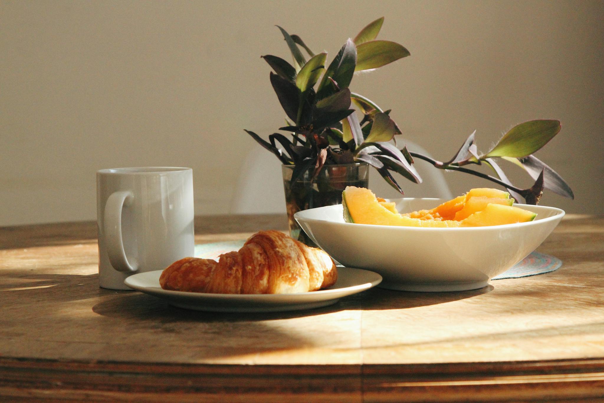 A warm and inviting breakfast setting with coffee, croissant, fresh fruit, and plant decor.